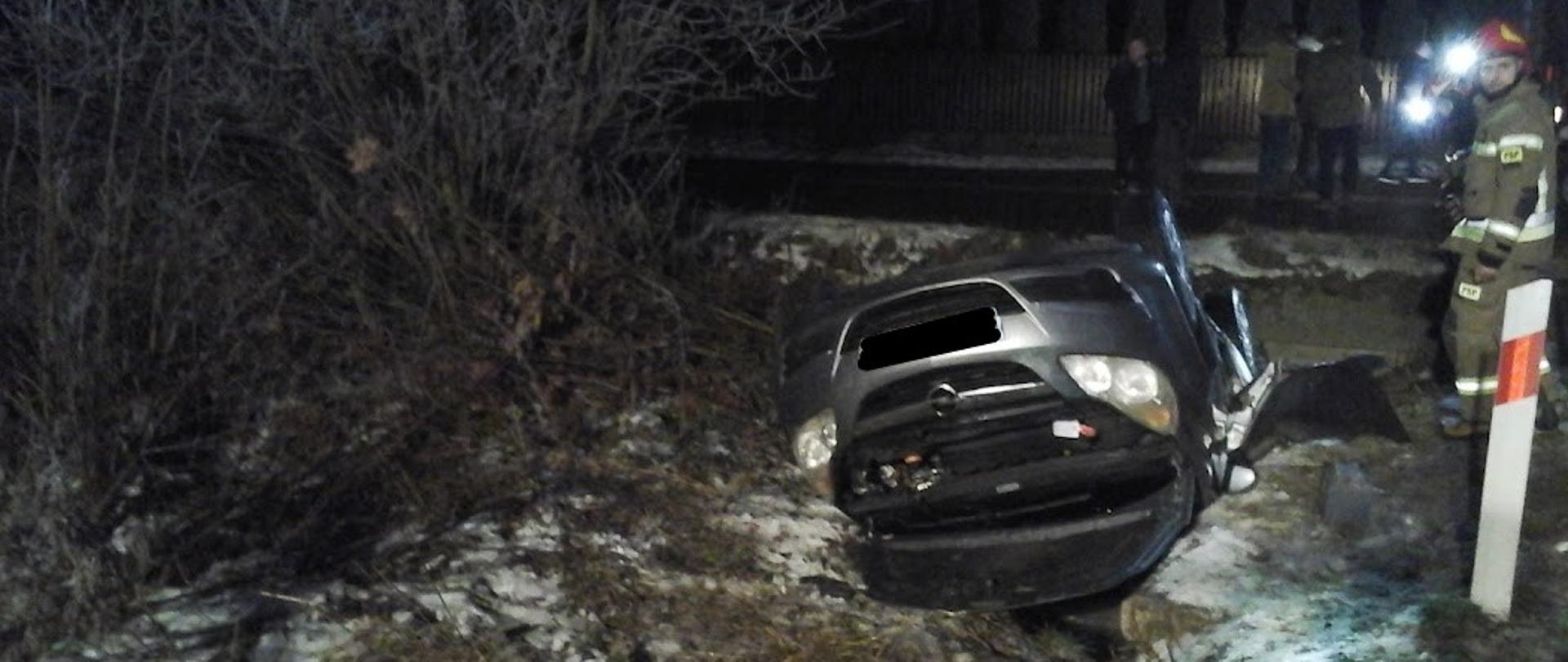 Zdjęcie przedstawia samochód osobowy leżący na dachu w rowie, obok funkcjonariusz Straży pożarnej 