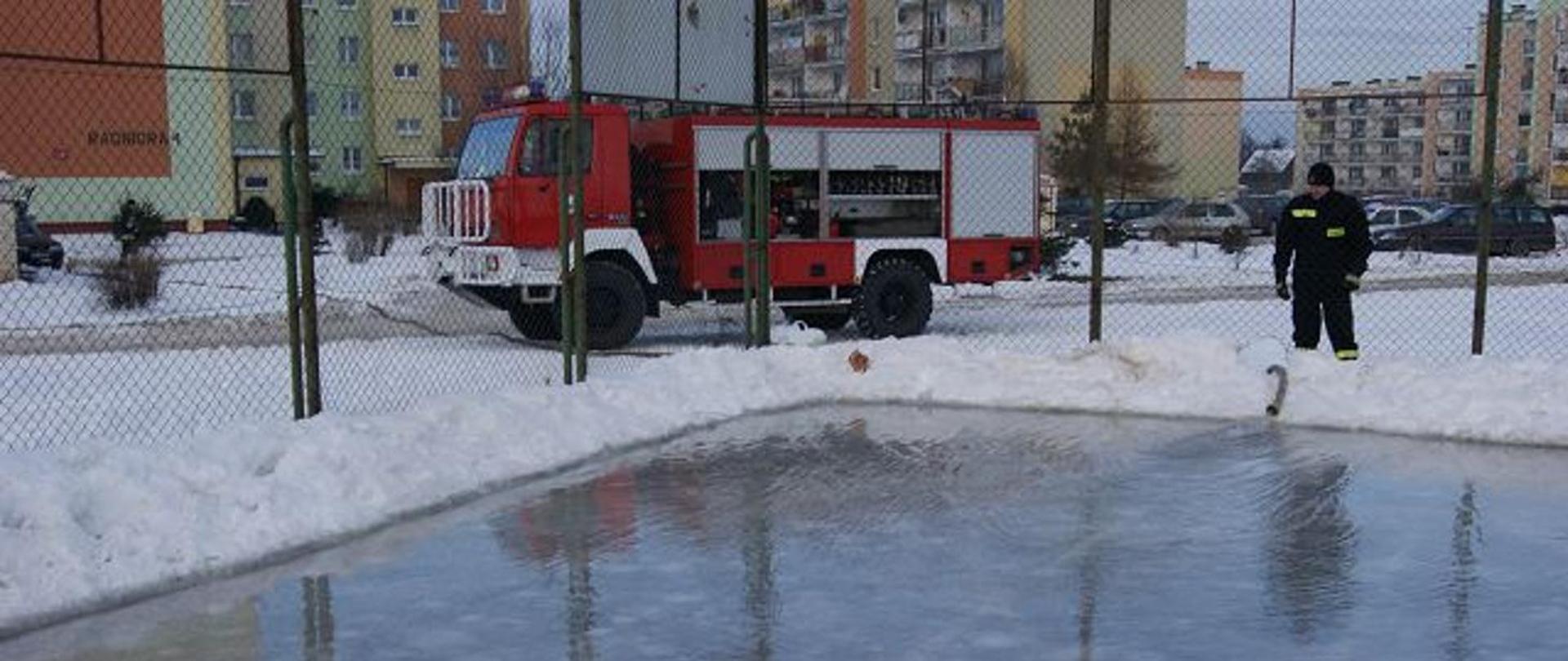 Obraz przedstawia strażaka przy lodowisku, w tle strażacki samochód