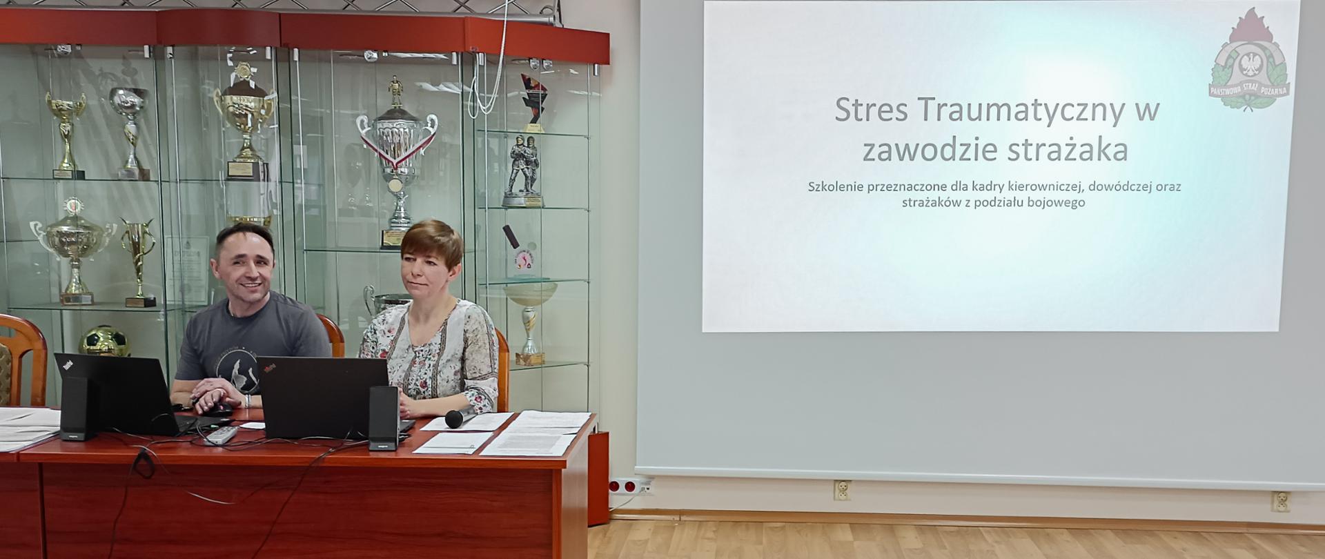 mężczyzna i kobieta siedzący przy biurku, za nimi przeszklona gablota z pucharami a po prawej ekran rzutnika z wyświetlonym slajdem na slajdzie napis "stres traumatyczny w zawodzie strażaka szkolenie przeznaczona dla kadry kierowniczej, dowódczej oraz strażaków z podziału bojowego"