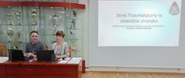 mężczyzna i kobieta siedzący przy biurku, za nimi przeszklona gablota z pucharami a po prawej ekran rzutnika z wyświetlonym slajdem na slajdzie napis "stres traumatyczny w zawodzie strażaka szkolenie przeznaczona dla kadry kierowniczej, dowódczej oraz strażaków z podziału bojowego"
