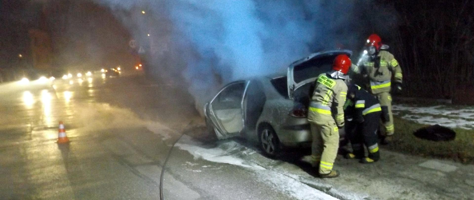Na zdjęciu widać trzech strażaków biorących udział w gaszeniu palącego się samochodu. W oddali unoszą się kłęby dymu.