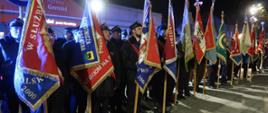 Narodowe Święto Niepodległości. Zdjęcie przedstawia plac przed pomnikiem Żołnierza Polskiego. Na tle budynków, w szeregu stoją poczty sztandarowe. Cztery pierwsze to poczty strażackie. Sztandarowi trzymają sztandary w pozycji pionowej. 