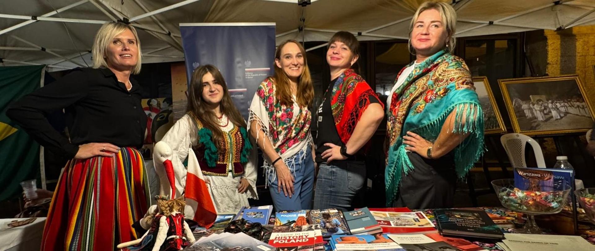 Ubrane w stroje ludowe kobiety stoją obok siebie wśród książek o Polsce 