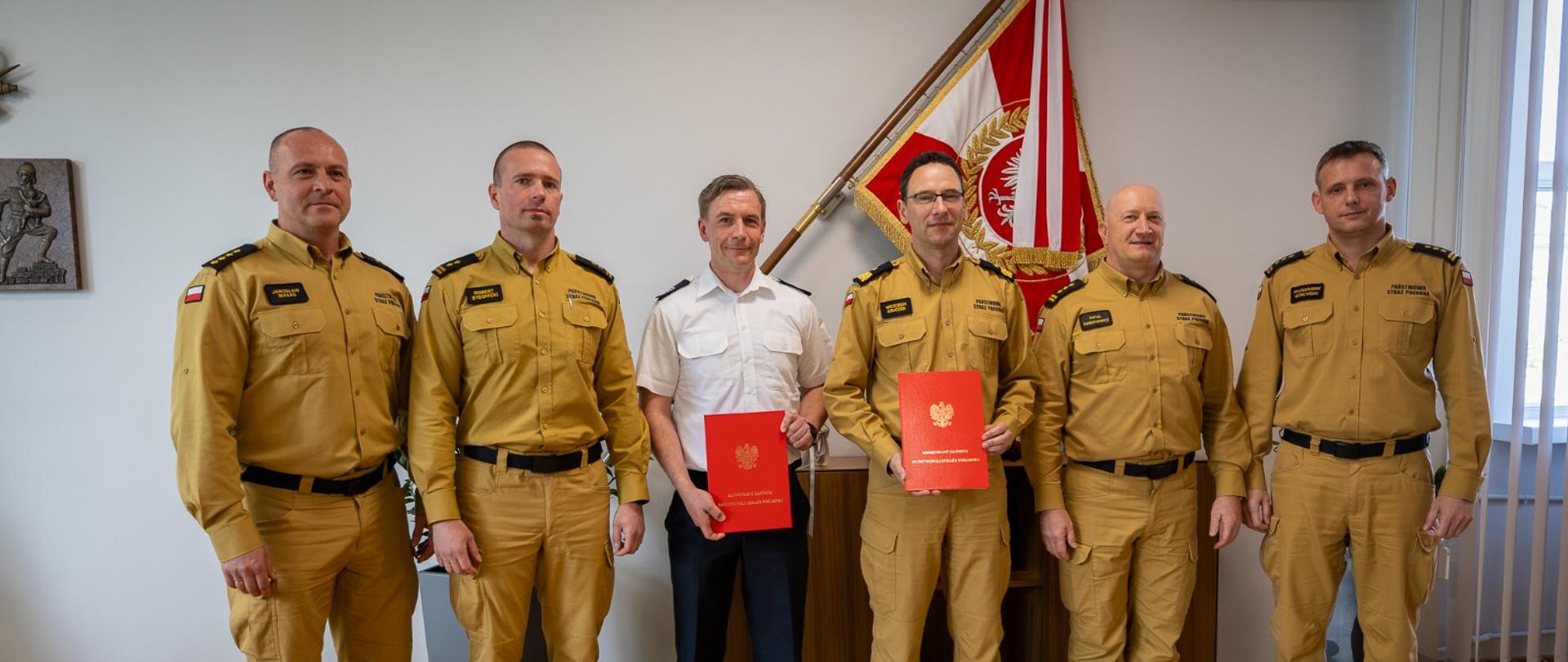 Grupa sześciu mężczyzn w mundurach PSP stoi w rzędzie. W środku Daniel Berger i nadbryg. Wojciech Kruczek trzymają czerwone teczki z godłem Polski. W tle znajduje się sztandar Państwowej Straży Pożarnej.