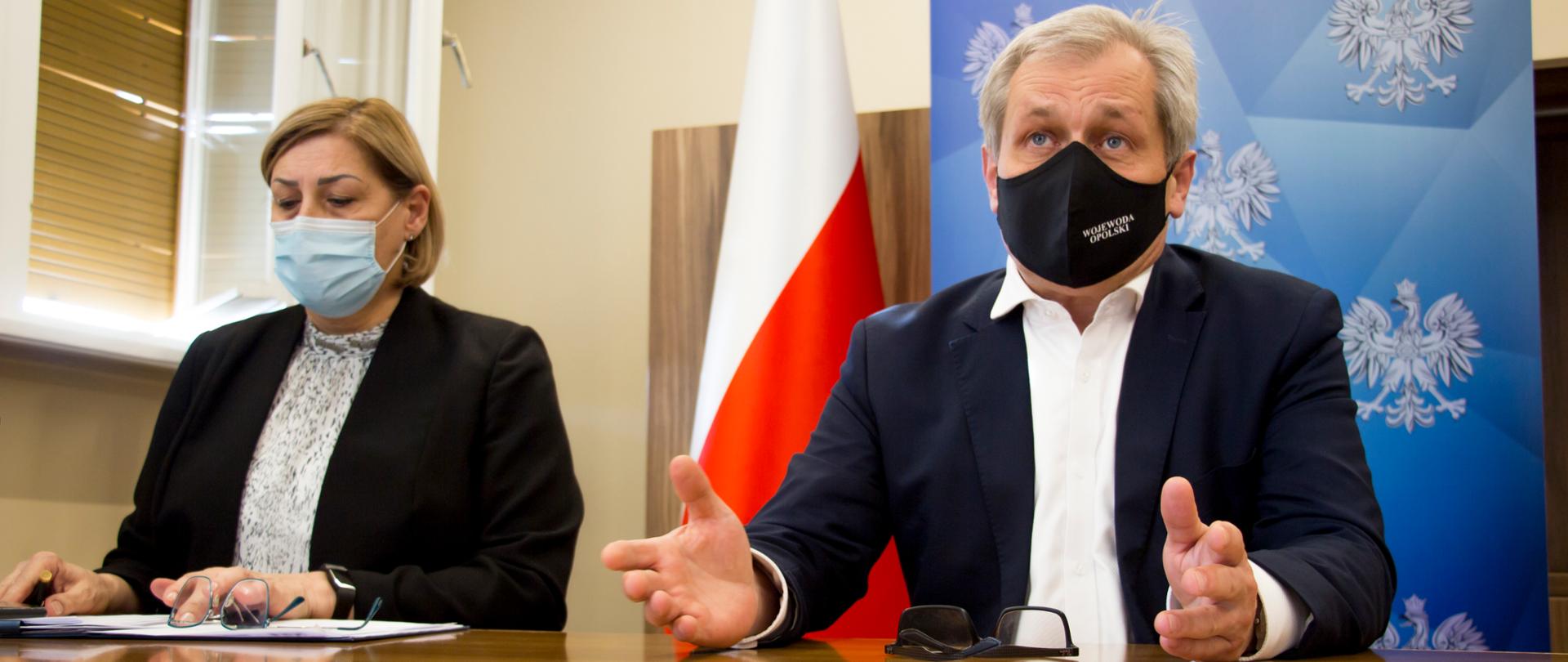 Na zdjęciu od prawej, siedzący za stołem wojewoda opolski Sławomir Kłosowski oraz - obok niego - z-ca dyrektora OOW NFZ Beata Cyganiuk. Wojewoda ma otwarte dłonie, spogląda przed siebie. Przed nim na stole leżą okulary. Dyrektor opolskiego NFZ, siedząca obok, ubrana w czarną marynarkę posgląda na notatki, jakie ma przed sobą. Obok niej na stole również lezą okulary. W tle meble oraz biało-czerwona flaga. 