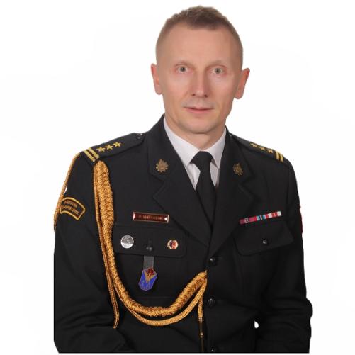 Na białym tle znajduje się strażak w mundurze wyjściowym. Mundur wyjściowy posiada przypięte odznaczenia, sznur galowy, baretki, odznaki oraz absolwentki. Strażak w stopniu starszego brygadiera patrzy przed siebie.