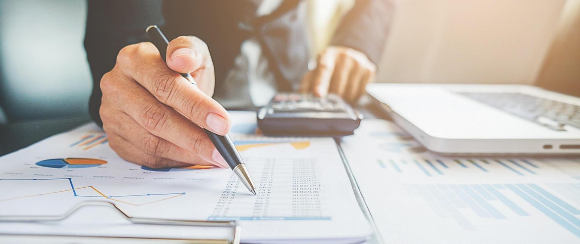 Męska dłoń z długopisem nad arkuszami z danymi liczbowymi i wykresami