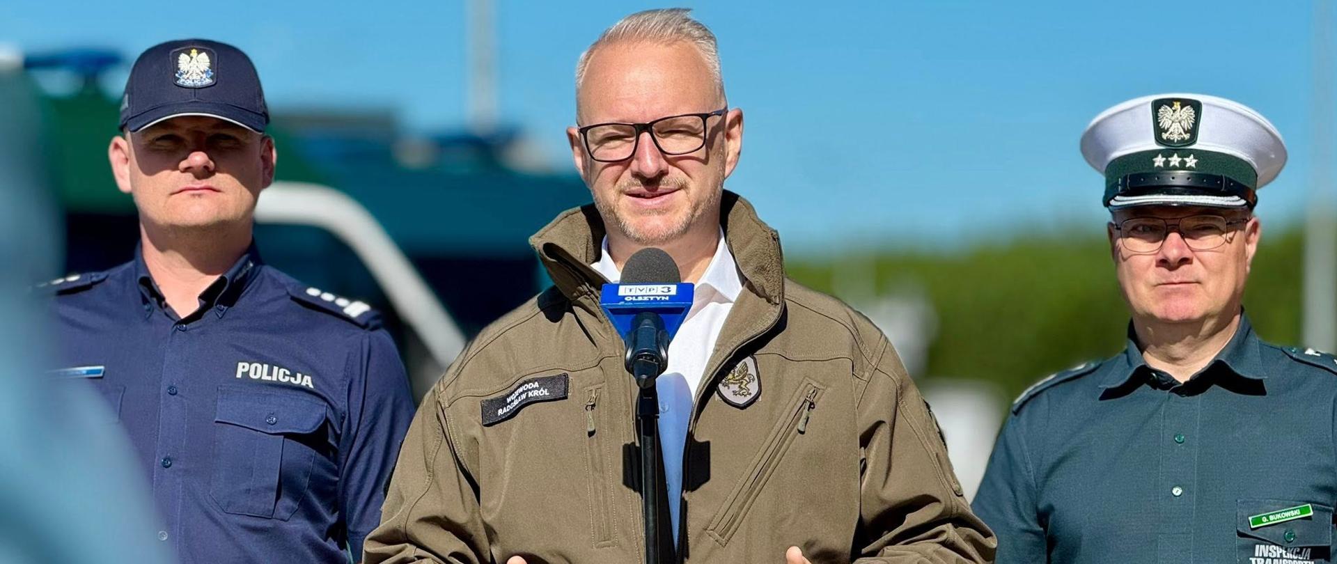 Trzech mężczyzn ubranych w mundurach stoi na tle radiowozów. Jeden z nich przemawia do stojącego przed nim mikrofonu. 