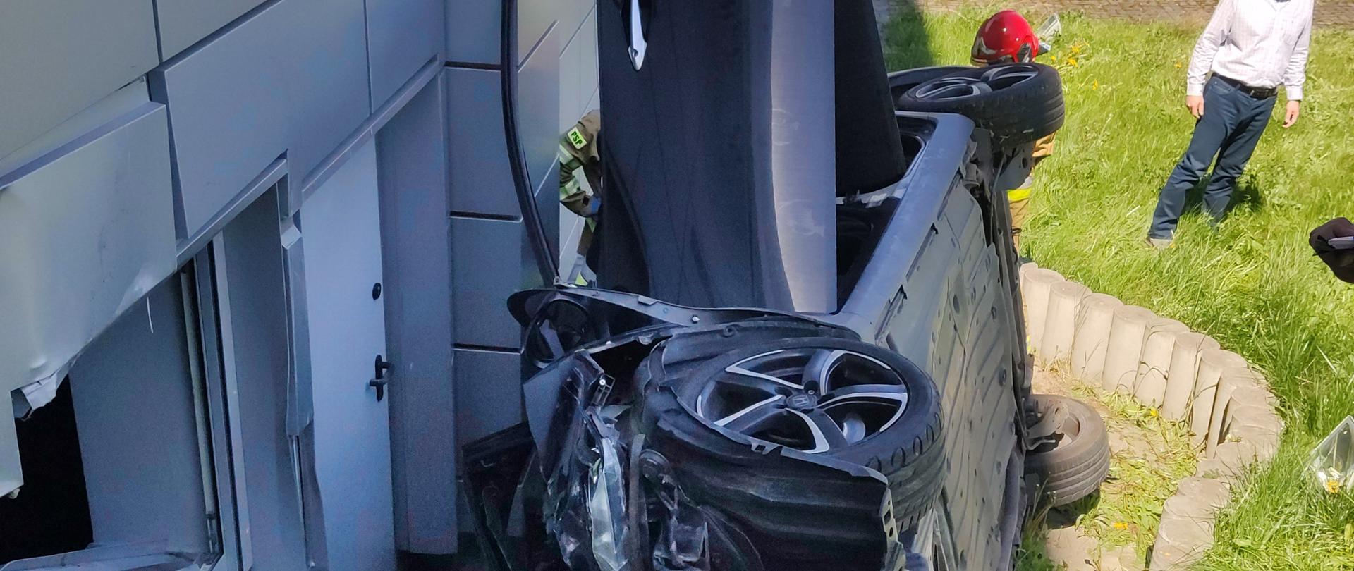 Zdjęcie przedstawia uszkodzony samochód osobowy ze zgniecioną maską i otwartymi drzwiami od strony kierowcy. Samochód leży na prawym boku tuz przy ścianie budynku. W tle widać strażaka oraz samochody strażackie.