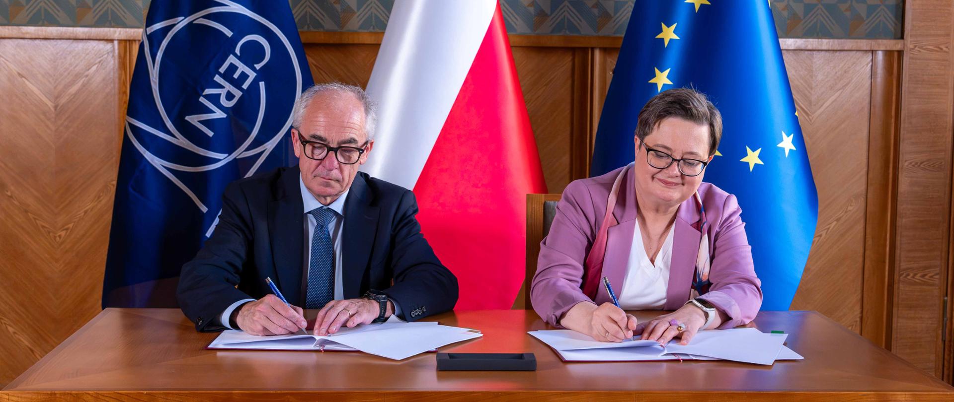 Dyrektor Generalny CERN Mark Thomson oraz wiceministra Katarzyna Lubnauer siedzą przy drewnianym stole i podpisują dokumenty. W tle stoją flagi CERN, Polski oraz Unii Europejskiej.
