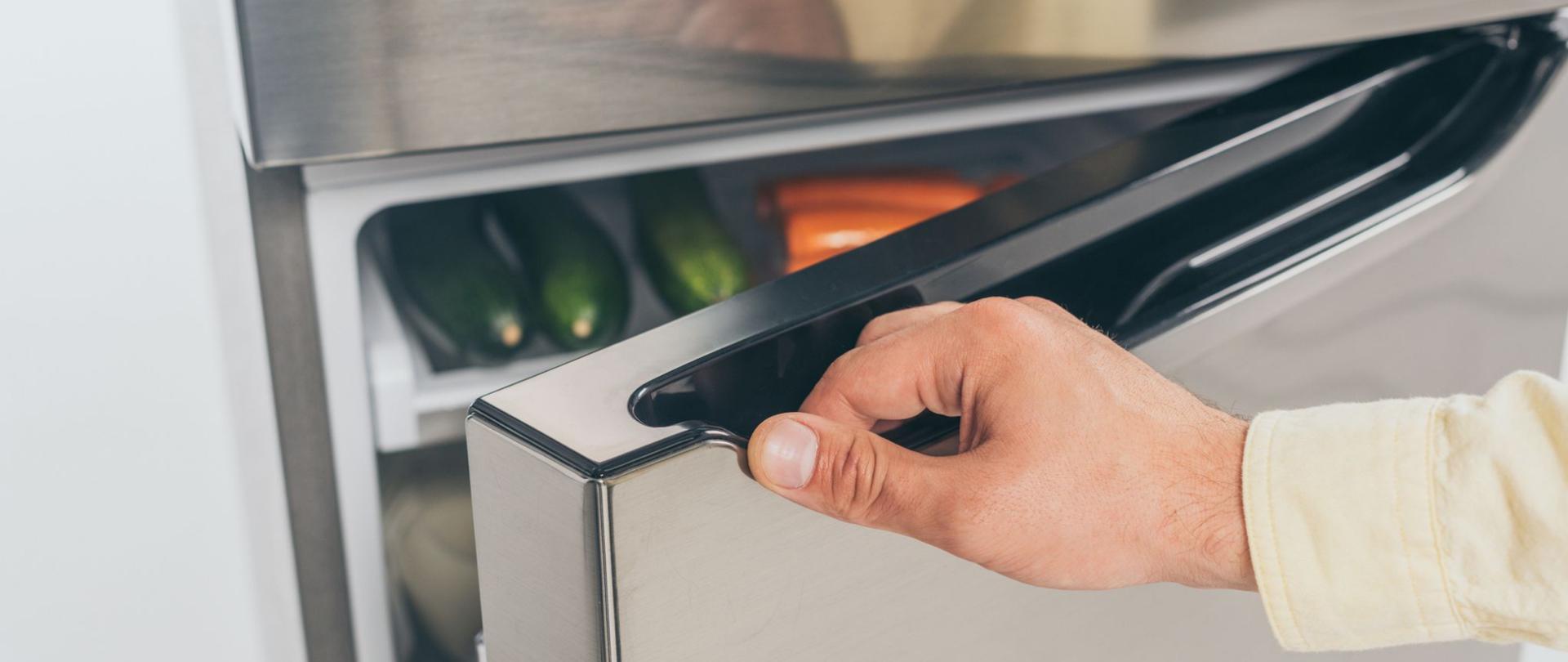 Na zdjęciu jest otwarta lodówka przez widoczną dłoń mężczyzny.