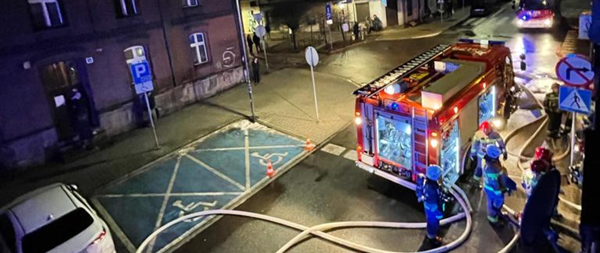 Zdjęcie przedstawia strażaków, wóz strażacki, rozwinięte linie wężowe, radiowóz policji. Ujęcie uchwycone z góry.