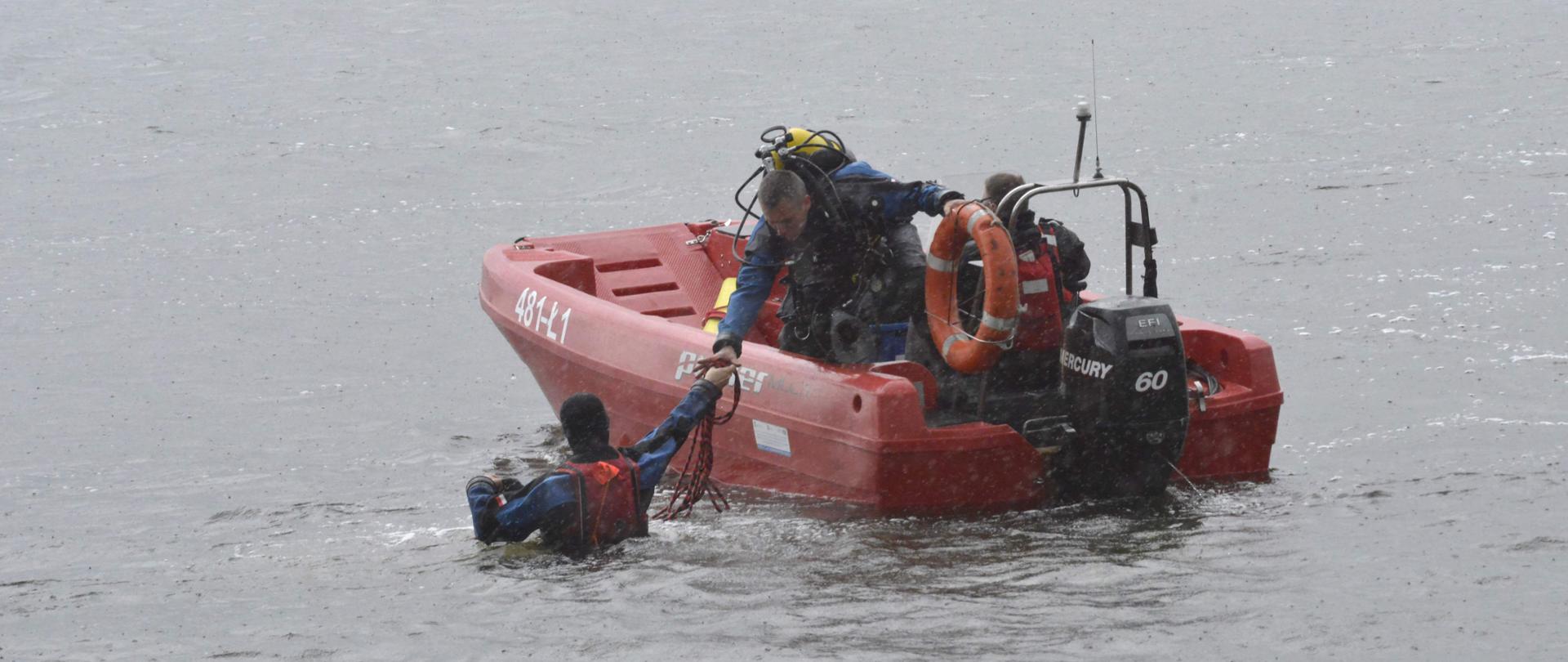 Na zdjęciu widać łódź ratowniczą Państwowej Straży Pożarnej, na jej pokładzie jeden ratownik a drugi ratownik w wodzie. 