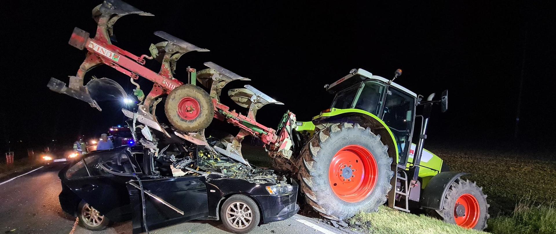 Zdjęcie przedstawia zderzenie samochodu osobowego z ciągnikiem rolniczym, który miał podczepiony pług. Samochód osobowy wjechał w tył ciągnika, a podczepiony osprzęt oparty jest na dachu pojazdu. 