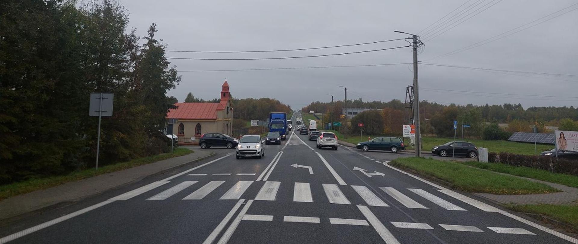Na zdjęciu widoczne jest skrzyżowanie drogi krajowej DK43 z ulicami Północną i Południową w miejscowości Lgota. Na pierwszym planie znajduje się przejście dla pieszych z wyraźnie oznakowanymi pasami. Na jezdni widoczne są strzałki kierunkowe – jeden pas umożliwia jazdę prosto i w prawo, natomiast drugi - jedynie w lewo. Na drodze porusza się kilka pojazdów, w tym samochody osobowe i ciężarówka. Po lewej stronie kadru znajduje się chodnik oraz kapliczka. Po prawej stronie widoczne są znaki drogowe, słup energetyczny oraz reklama. W tle rozciąga się krajobraz z drzewami w jesiennych barwach pod pochmurnym, szarym niebem.