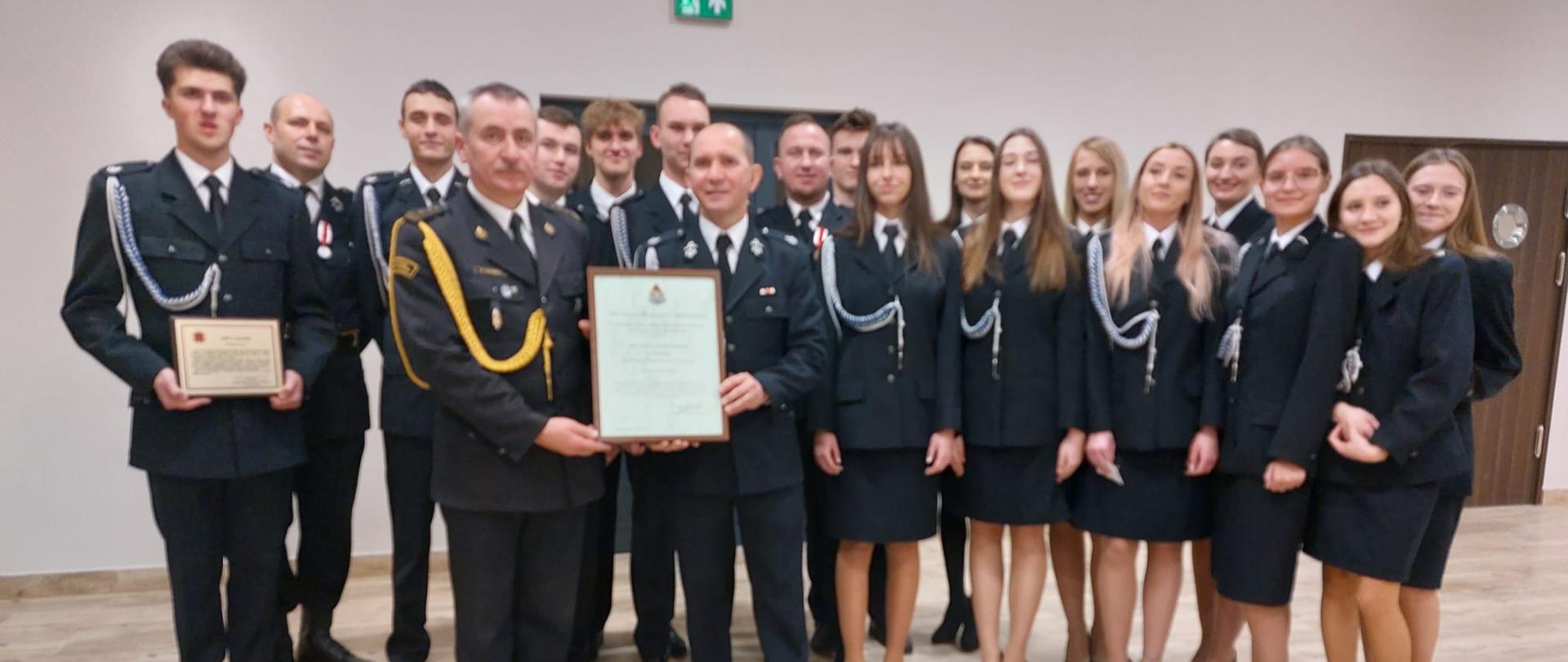 Dwóch strażaków trzyma oprawiony w ramkę dokument. Za nimi stoją strażacy ustawieni w półkole. Strażak stojący jak pierwszy z lewej strony trzyma w rękach pamiątkowa tabliczkę. Wszyscy znajdują się w pomieszczeniu z jasnymi ścianami i jasna podłogą.