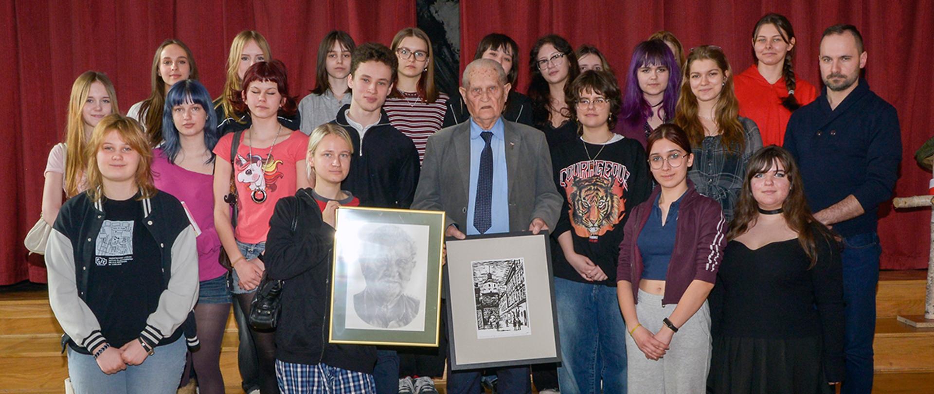 Grupowe zdjęcie na scenie z czerwonymi kotarami w tle. Pośrodku stoi starszy mężczyzna (F. Szopa) w garniturze, trzymający dwie oprawione prace graficzne. Wokół niego ustawiona jest grupa kilkunastu młodych osób w różnorodnych, codziennych strojach. Wszyscy stoją na drewnianej podłodze i schodkach sceny, patrząc w stronę aparatu.