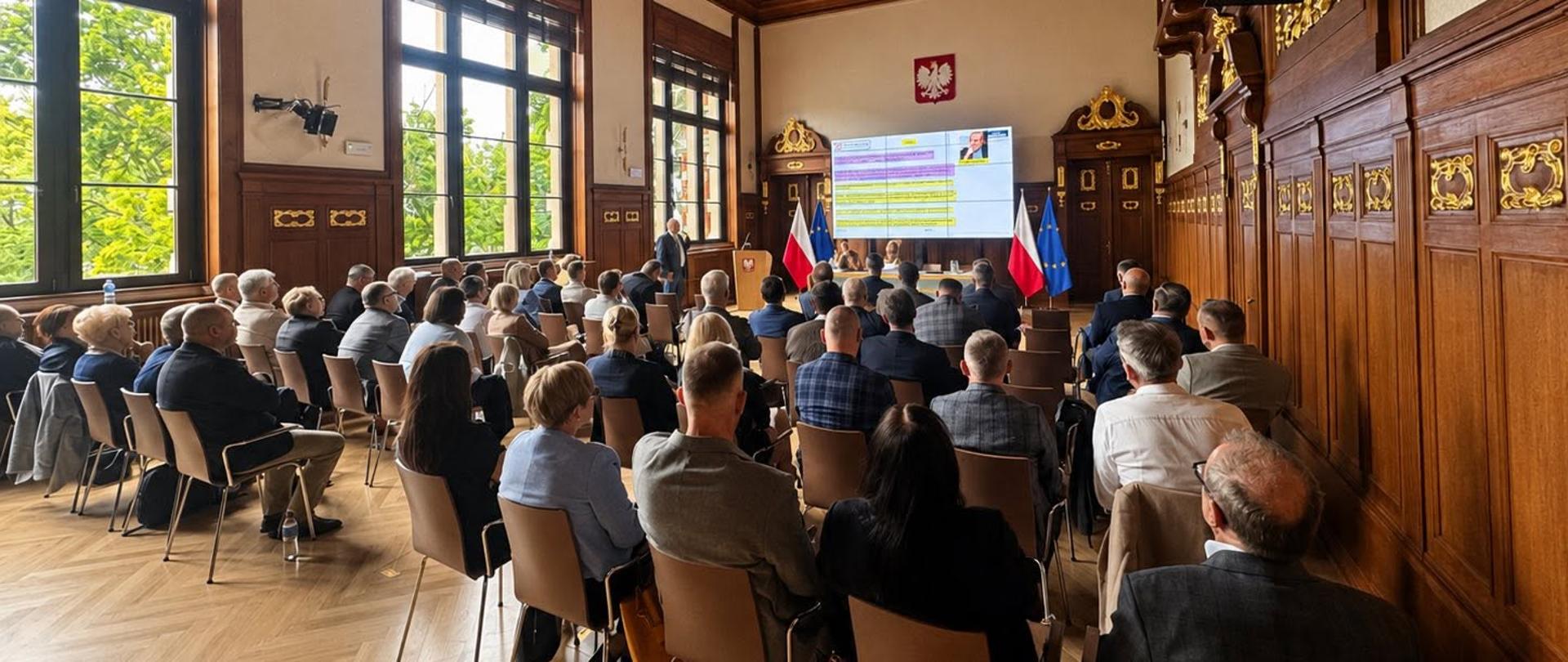 W dużej, drewnianej sali konferencyjnej uczestnicy siedzą i słuchają prezentacji. Na froncie sali widoczny jest ekran, a obok niego flagi Polski i Unii Europejskiej. Przez wysokie okna wpada naturalne światło.