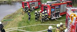 Zdjęcie przedstawia wozy pożarnicze oraz strażaków podczas ćwiczeń. w tle zbiornik wodny, z którego jest czerpana woda.