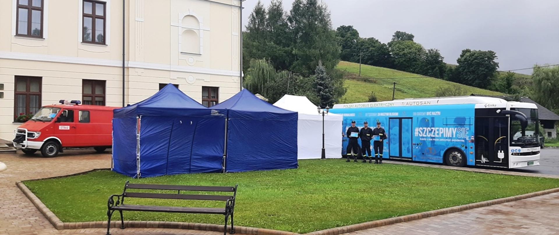 Kolorowe zdjęcie wykonane w przestrzeni otwartej w dzień. Na pierwszym planie zdjęcia widoczny zielony skwer i ławka. Na środkowym planie białe i niebieskie pawilony namiotowe, autobus w niebieskim kolorze z Widocznym białym napisem Szczepimy Się. Przed autobusem stoją trzej umundurowanych w ubrania koszarowe strażacy w czapkach na głowach i trzymają materiały informacyjne. Po lewej stronie obok namiotów stoi czerwony samochód pożarniczy z napisem Straż.Z tyłu widoczny fragment dużego budynku i zielone wzgórze z drzewami.
