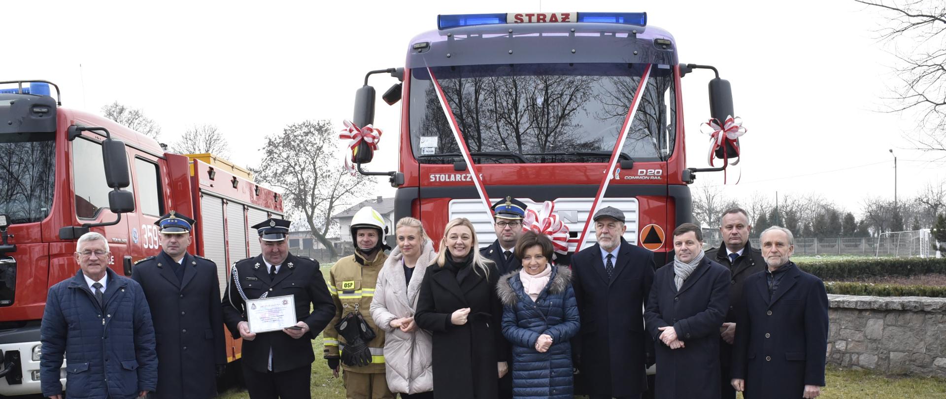 grupa kobiet i mężczyzn stoi przed samochodem strażakim, przód auta jest ozdobiony biało czerrwoną szarfą