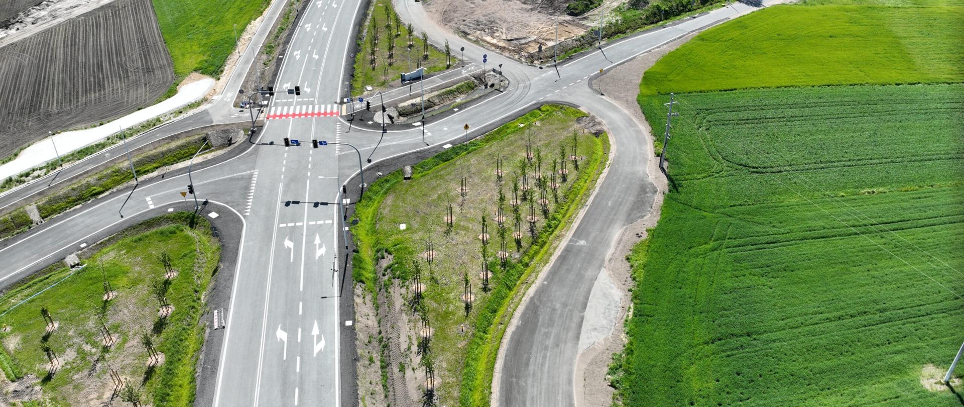 Widok z lotu ptaka na nowo wybudowane skrzyżowanie dróg na terenach wiejskich. Przez środek kadru przebiega szeroka jezdnia z oznakowaniem poziomym, przejściem dla pieszych i sygnalizacją świetlną. Od głównej drogi odchodzą drogi lokalne i łącznice. Wokół znajdują się zielone pola uprawne, pojedyncze zabudowania gospodarcze oraz niewielkie zadrzewienia. Widoczne są także tereny po robotach ziemnych i młode nasadzenia drzew przy drodze. Zdjęcie wykonano w słoneczny dzień.