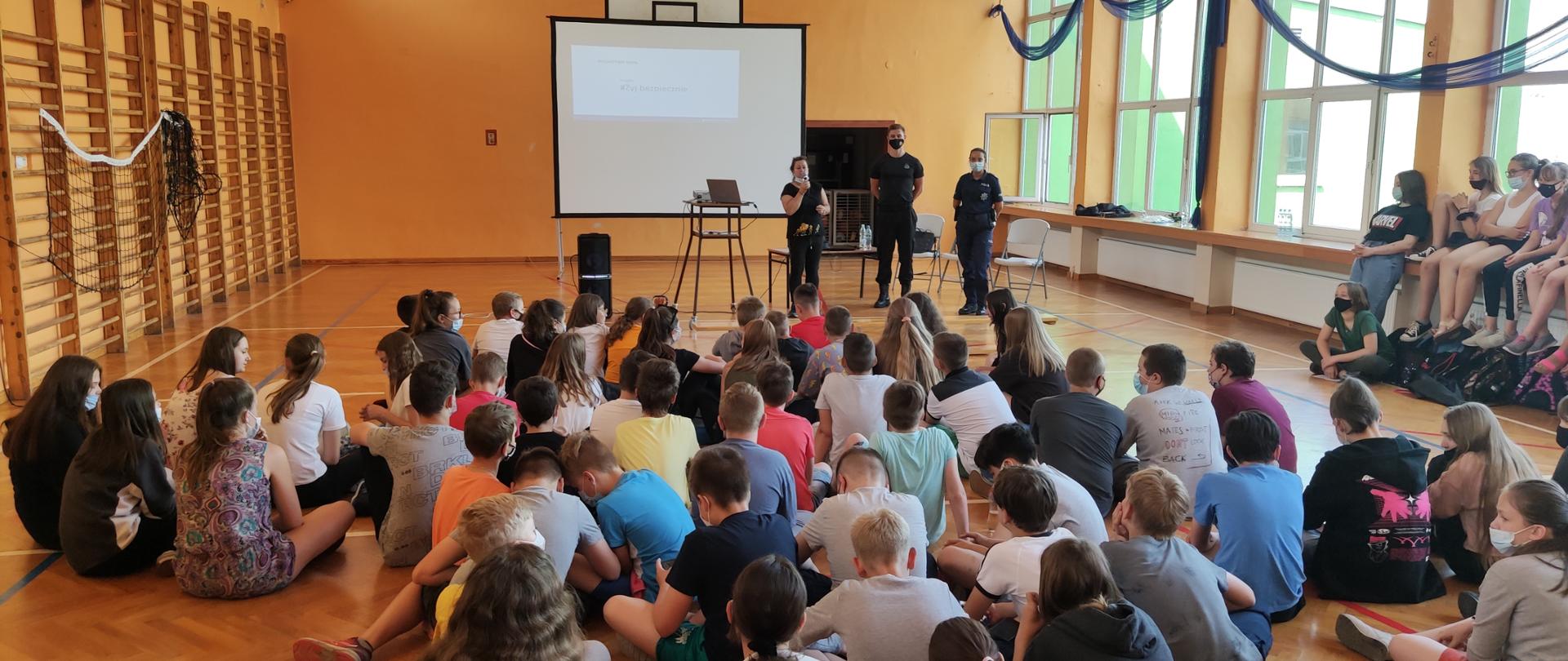 Zdjęcie przedstawia prowadzących prelekcje edukacyjną. W tle ekran rzutnika. Zdjęcie z sali gimnastycznej, na której zgromadziły się przed przemawiającymi dzieci z MZS Radziejów wraz z nauczycielami. 