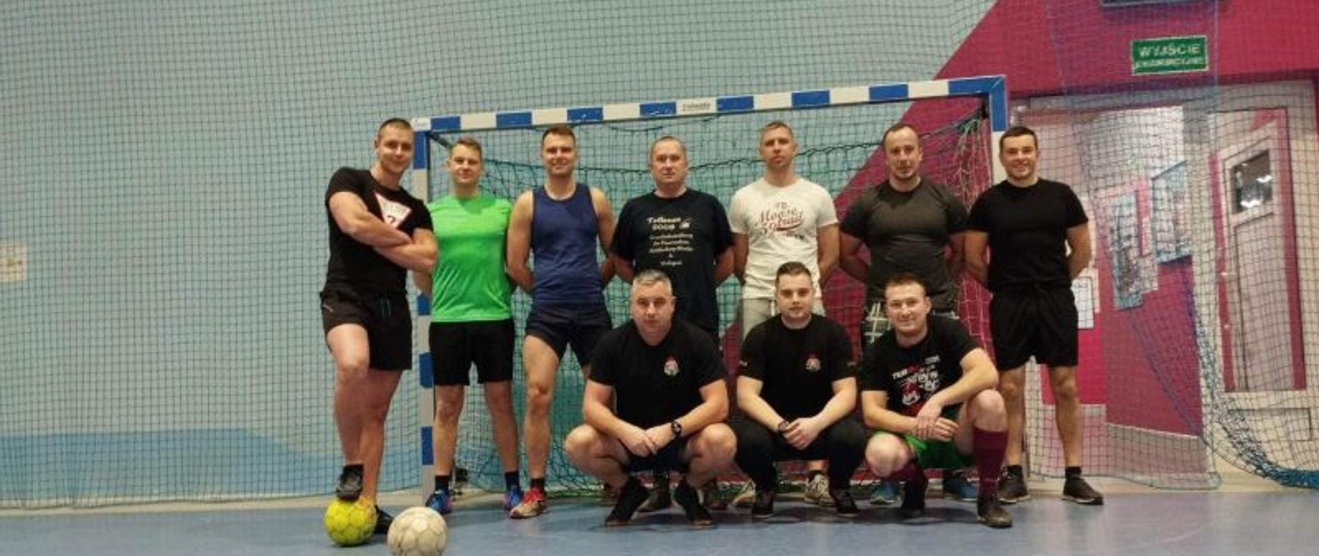 Centralny punkt zdjęcia stanowią strażacy ubrany w sportowe stroje. Strażacy stoją w bramce. W tle hala sportowa