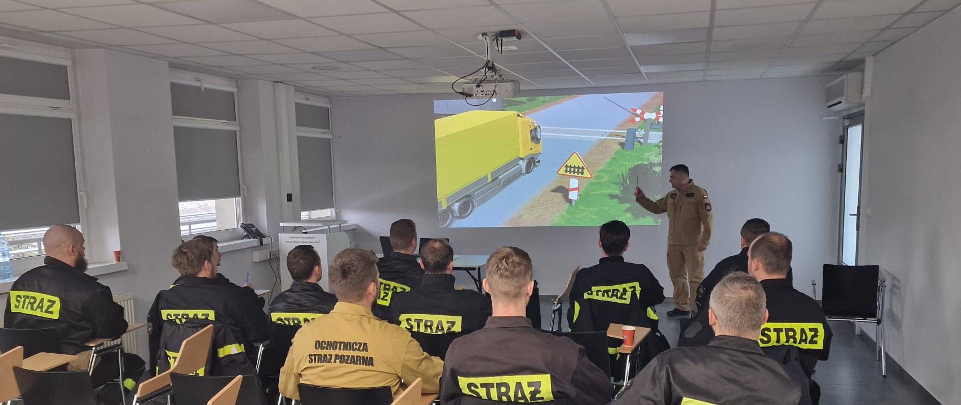 Na zdjęciu widać salę szkoleniową, w której kilkunastu strażaków Ochotniczej Straży Pożarnej siedzi na krzesłach, ustawionych w kilku rzędach. Wszyscy są zwróceni w stronę instruktora. Strażacy mają na sobie czarne lub piaskowe ubrania służbowe z dużym, żółtym napisem „STRAŻ” na plecach. Z przodu sali stoi prowadzący szkolenie strażak w piaskowym mundurze, który gestem ręki wskazuje na wyświetlaną prezentację. Na ścianie widoczny jest slajd z ilustracją ciężarówki stojącej przy przejeździe kolejowym oraz znakami ostrzegawczymi. Nad salą zamontowany jest projektor. 
