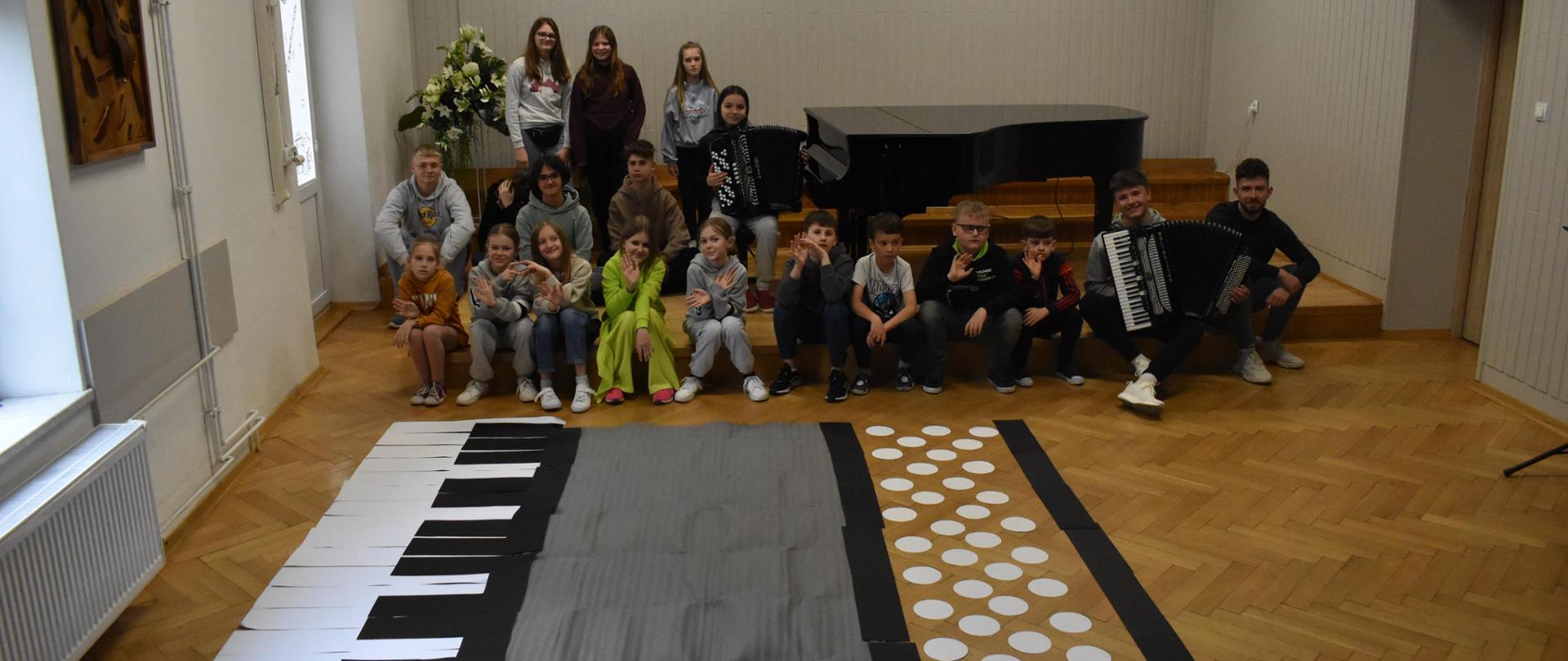 na zdjęciu, na podłodze auli PSM w Jaśle, na pierwszym planie widnieje wizerunek dużego akordeonu, wykonany z tekturowych biało-czarnych elementów oraz szarego materiału. Na drugim planie widoczni są uczniowie, którzy siedzą lub stoją w trzech rzędach. Dwoje uczniów trzyma na swoich kolanach akordeon. Kolorystyka zdjęcia jest biało-szaro-czarno-brązowo-zielona.