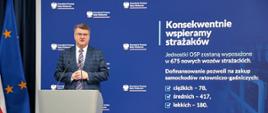 Minister Maciej Wąsik przemawiający zza mównicy na tle prezentacji multimedialnej