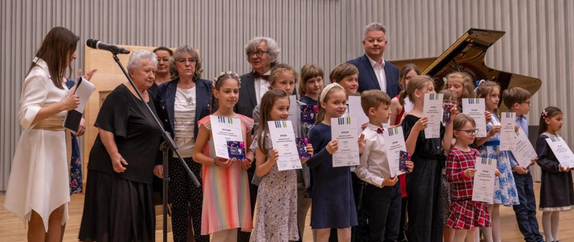 Zdjęcie przedstawiające laureatów Ogólnopolskiego "Łowieckiego" Konkursu Miniatur Fortepianowych we Wrocławiu. Laureaci stoją z dyplomami na scenie w towarzystwie jurorów.