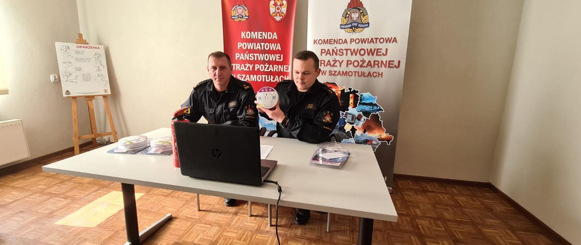 Dwóch strażaków w mundurach prezentuje czujkę podczas wideokonferencji.