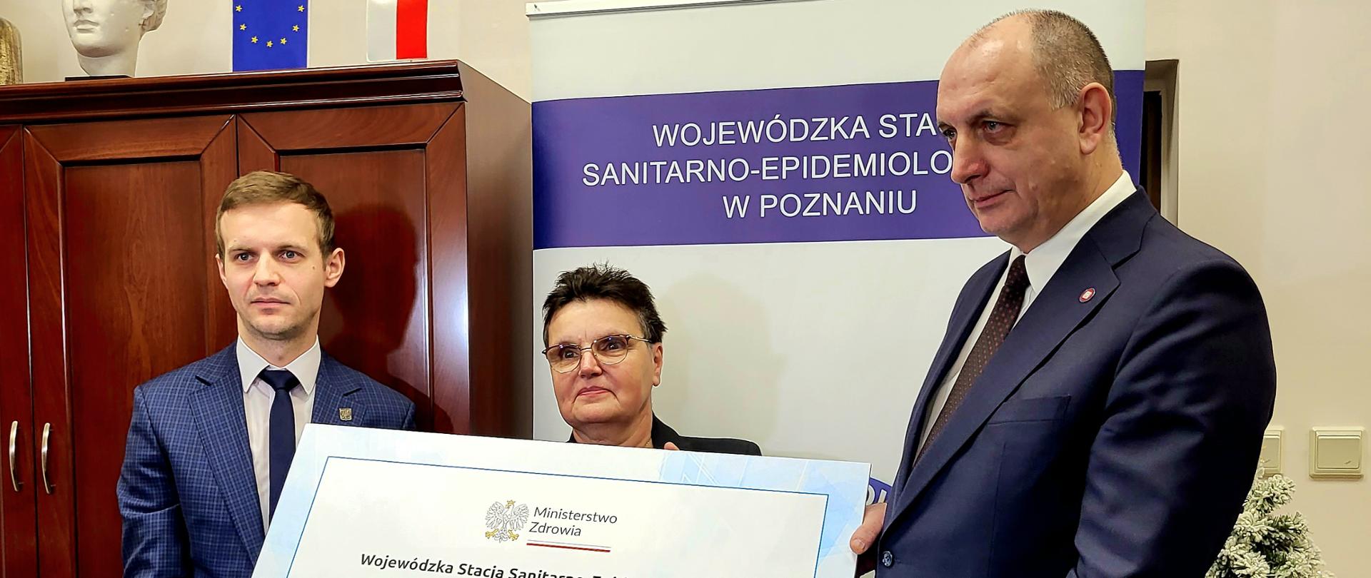 Wiceminister zdrowia Marcin Martyniak, dyrektor WSSE Poznań Jadwiga Kuczma-Napierała, Główny Inspektor Sanitarny wz. Krzysztof Saczka trzymają wielki czek z kwotą 6,9 mln zł. 