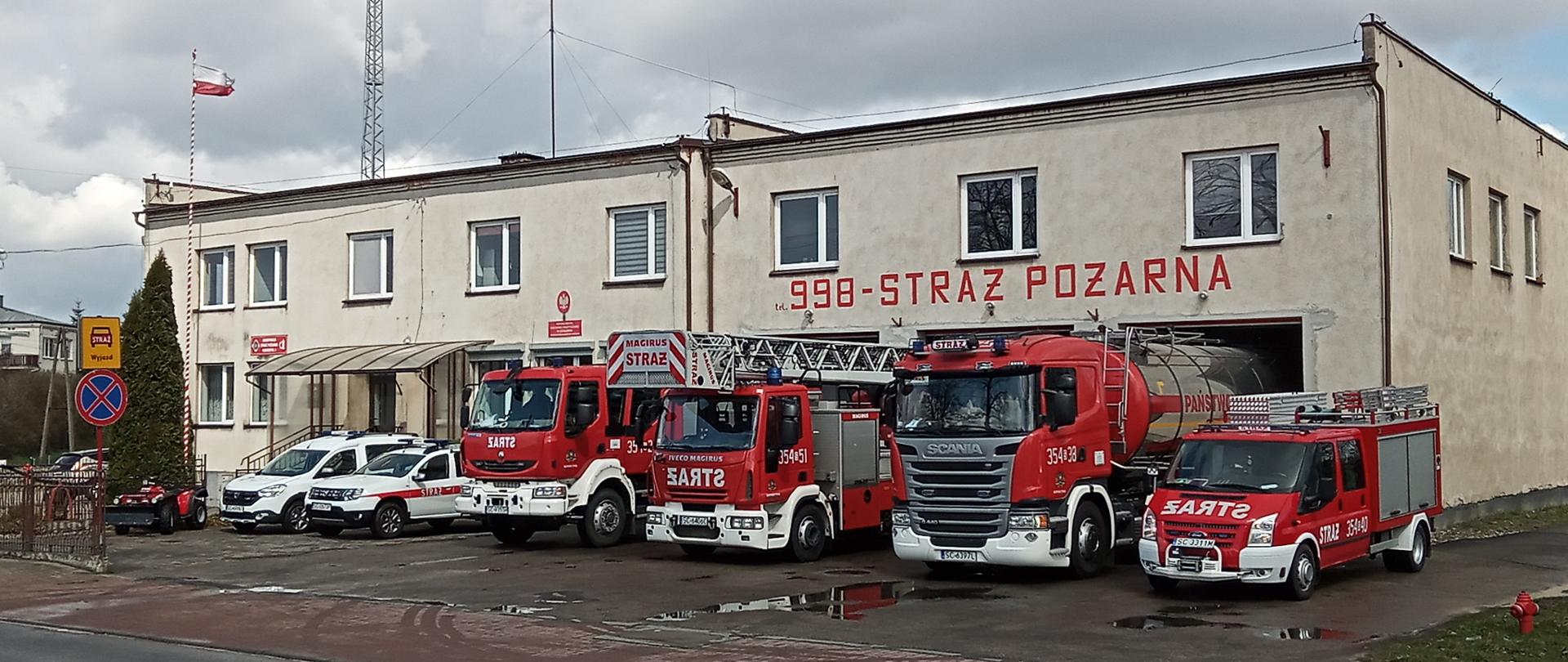 Zdjęcie przedstawia Jednostkę Ratowniczo Gaśniczą nr 4 w Koniecpolu. Na zdjęciu widoczny obiekt Jednostki Ratowniczo Gaśniczej przed obiektem znajdują się pojazdy będące na wyposażeniu jednostki.