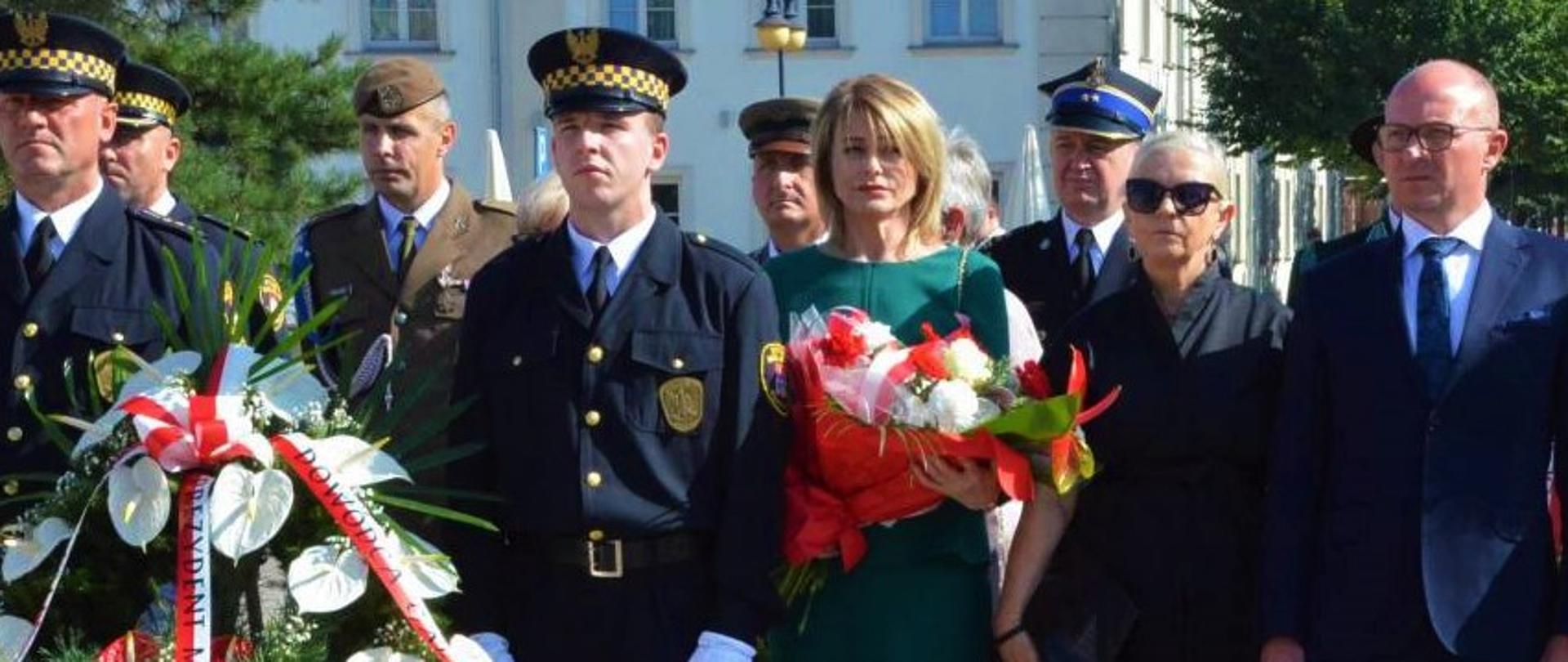 Zgromadzenie przed pomnikiem w celu złożenia kwiatów. Na zdjęciu kilkanaście osób z wieńcem okolicznościowym. Wśród nich komendant miejski PSP.