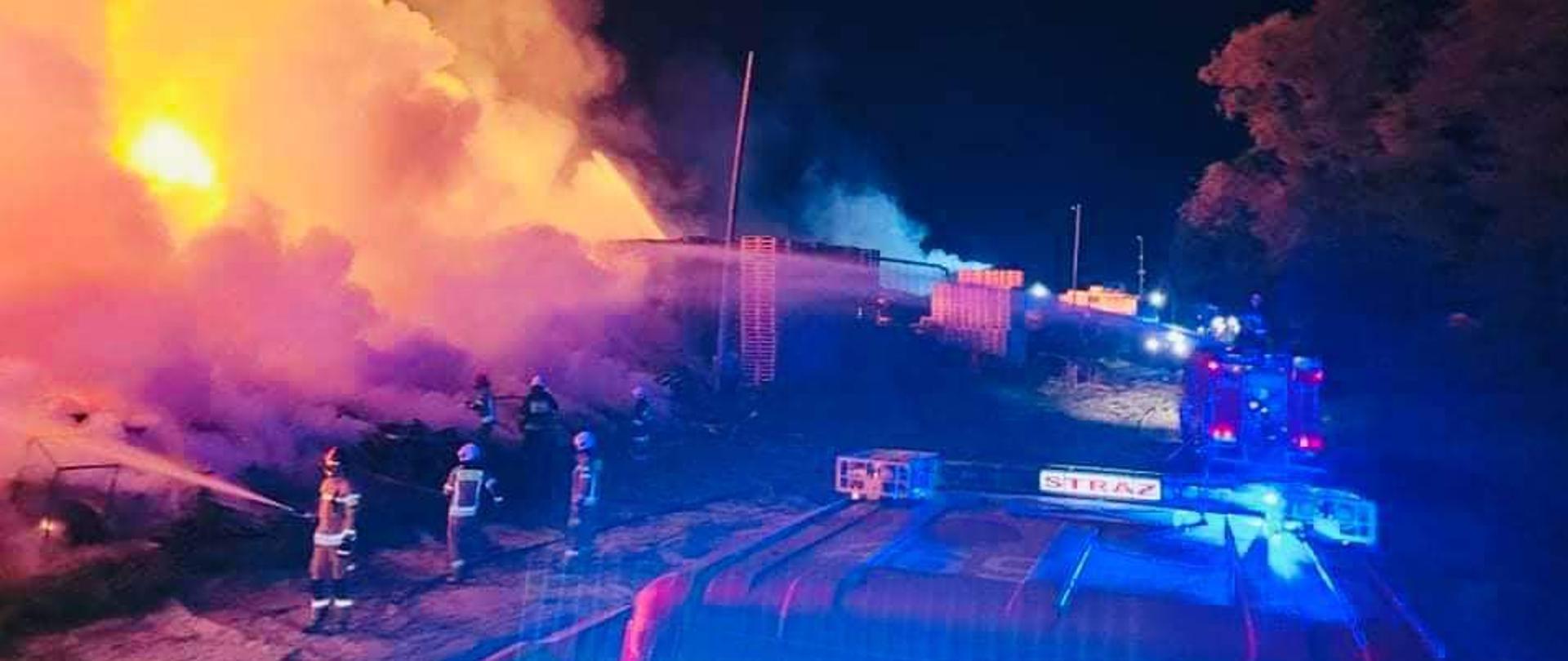 strażacy gaszą duży pożar, w tle pojazdy pozarnicze, dużo dymu i ogień