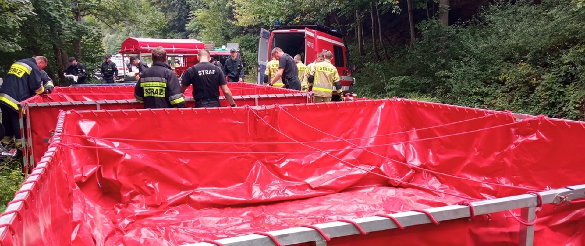 Na zdjęciu widoczni strażacy w czarnych i piaskowych ubraniach koszarowych i bojowych podczas ćwiczeń i rozkładnia zbiorników na wodę typu tanker o pojemności 13 m3. Na wprost widoczne dwa czerwone zbiorniki i samochody pożarnicze po obu stronach drogi las iglasty.