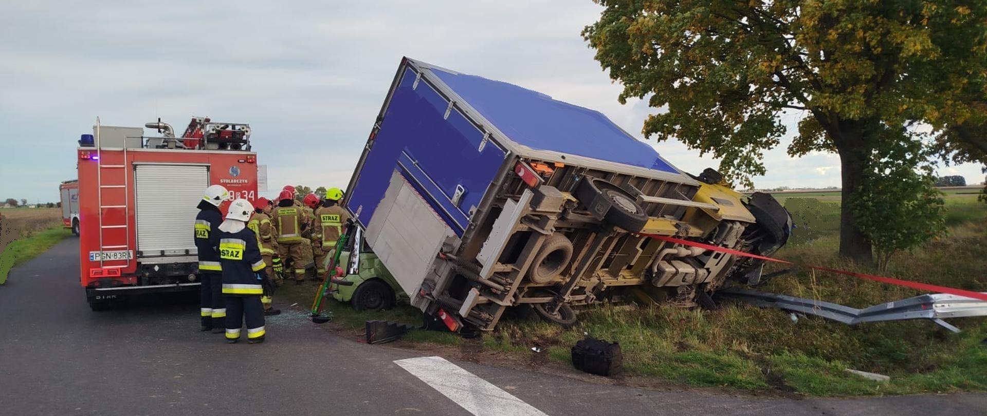 Na zdjęciu widać tył oraz prawy bok samochodu dostawczego Pojazd dostawczy ustabilizowany jest za pomocą podpory oraz pasa ściągającego. Po lewej stronie zdjęcia widać wozy strażackie oraz strażaków obsługujących to zdarzenie.