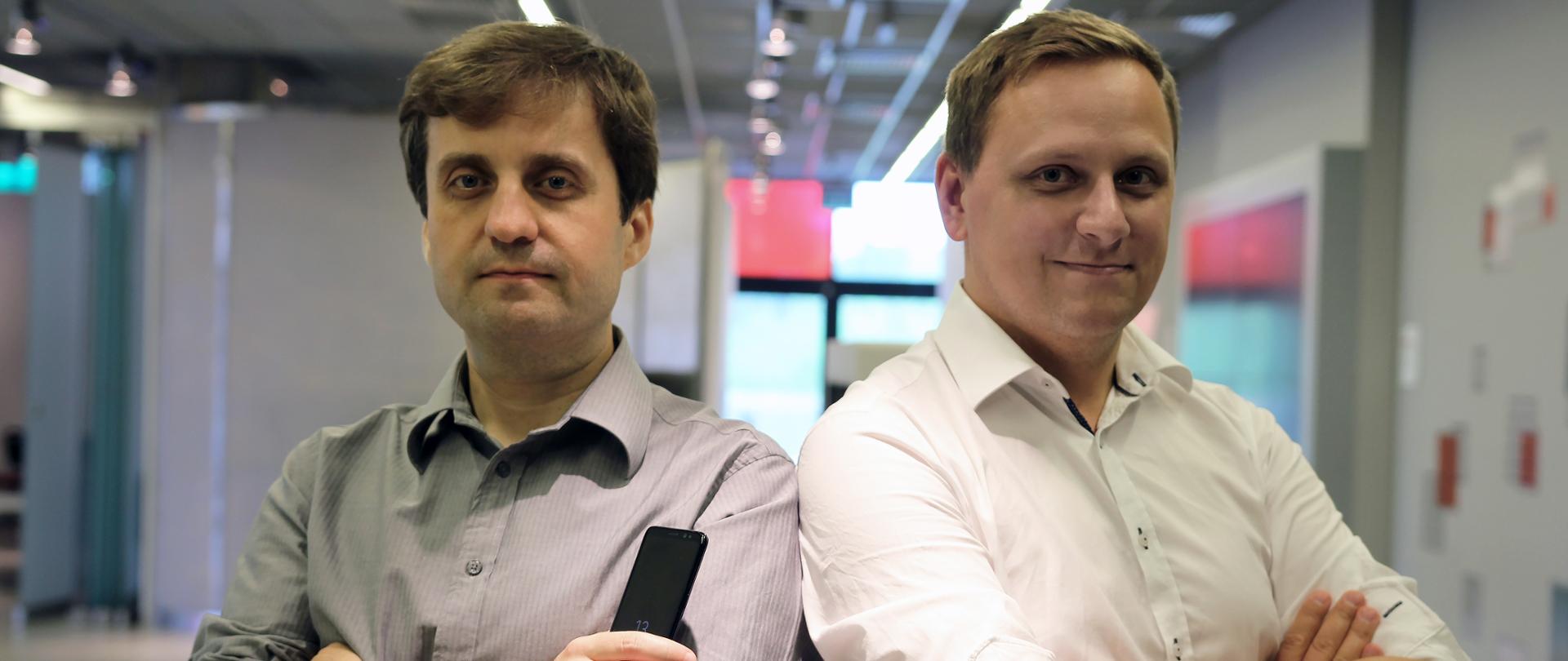 W ciasnym kadrze od pasa w górę dwóch mężczyzn w wieku około 30 lat, w jasnych koszulach, na tle dużej jasnej sali. Każdy z mężczyzn z rękami założonymi jedna na drugą. Mężczyzna z prawej strony trzyma w prawej dłoni czarny telefon komórkowy.