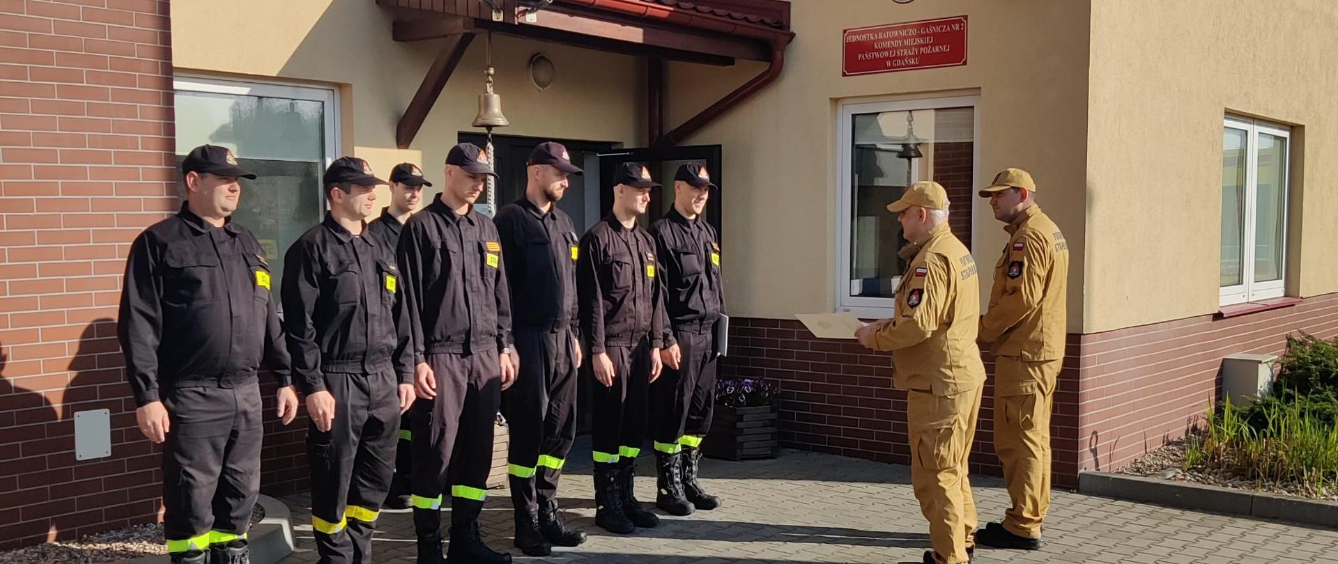 Strażacy stoją w szeregu przez budynkiem JRG 2. Naprzeciw komendant miejski PSP.