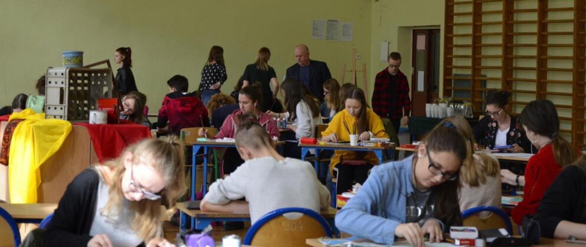 Sala gimnastyczna, na pierwszym i drugim planie przy stolikach młodzież pracuje nad pracami plastycznymi, mają rozłożone farby i kredki, po lewej stronie ustawiona martwa natura. Na ostatnim planie osoby wchodzące i wychodzący z sali gimnastycznej