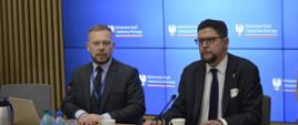 Za stołem siedzi wiceminister Szeptycki i mężczyzna w garniturze, za nimi wbudowany w ścianę wielki ekran wyświetlający logo MNiSW.