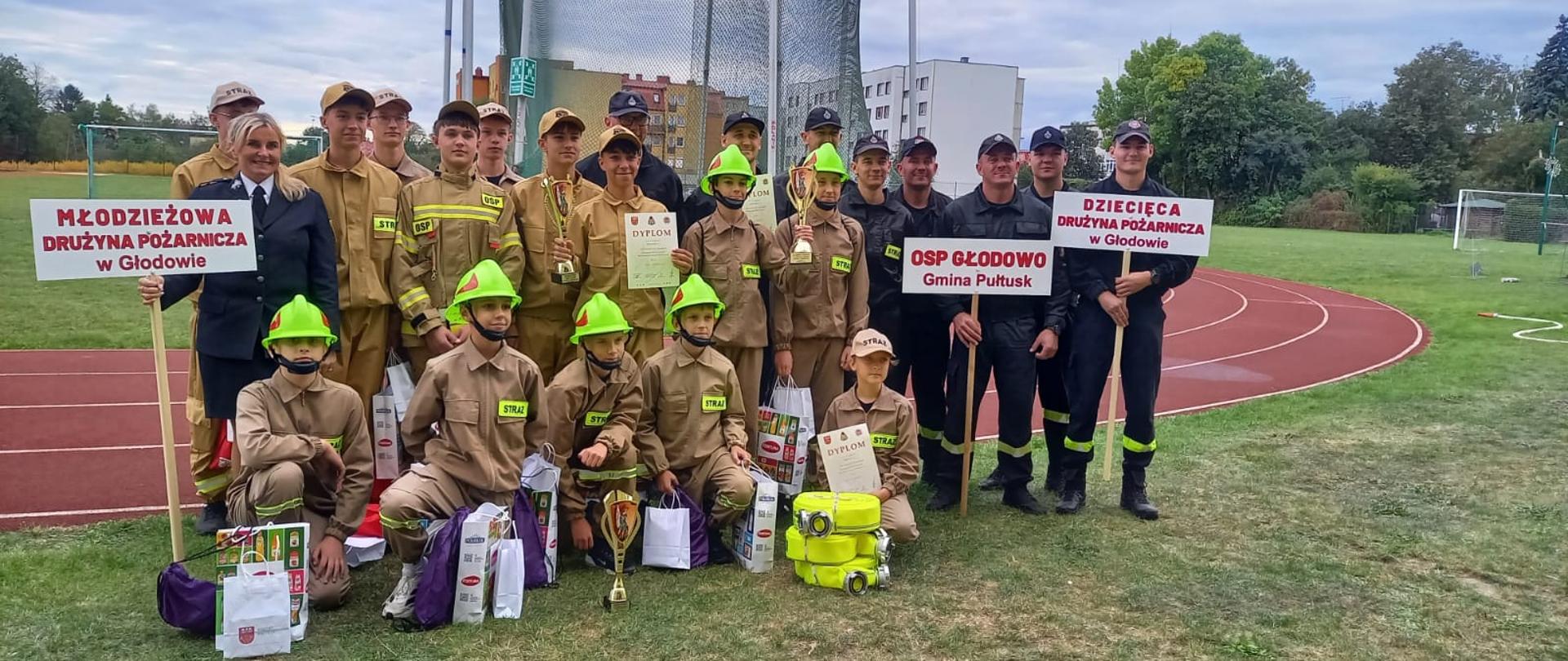 Na zdjęciu grupa osób w różnym wieku pozuje na boisku lekkoatletycznym. Widać dwie drużyny w strojach strażackich – dzieci w jasnobrązowych mundurach i jasnozielonych kaskach oraz starszą młodzież w podobnych ubraniach, ale bez kasków. Z boku stoją także dorośli w czarnych mundurach strażackich. Dwie osoby trzymają tabliczki z napisami: „Młodzieżowa Drużyna Pożarnicza w Głodowie” oraz „Dziecięca Drużyna Pożarnicza w Głodowie”. Przed grupą leżą nagrody – puchary, dyplomy i torby z upominkami. W tle widać bieżnię, fragment boiska, budynki mieszkalne i drzewa.