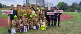 Na zdjęciu grupa osób w różnym wieku pozuje na boisku lekkoatletycznym. Widać dwie drużyny w strojach strażackich – dzieci w jasnobrązowych mundurach i jasnozielonych kaskach oraz starszą młodzież w podobnych ubraniach, ale bez kasków. Z boku stoją także dorośli w czarnych mundurach strażackich. Dwie osoby trzymają tabliczki z napisami: „Młodzieżowa Drużyna Pożarnicza w Głodowie” oraz „Dziecięca Drużyna Pożarnicza w Głodowie”. Przed grupą leżą nagrody – puchary, dyplomy i torby z upominkami. W tle widać bieżnię, fragment boiska, budynki mieszkalne i drzewa.