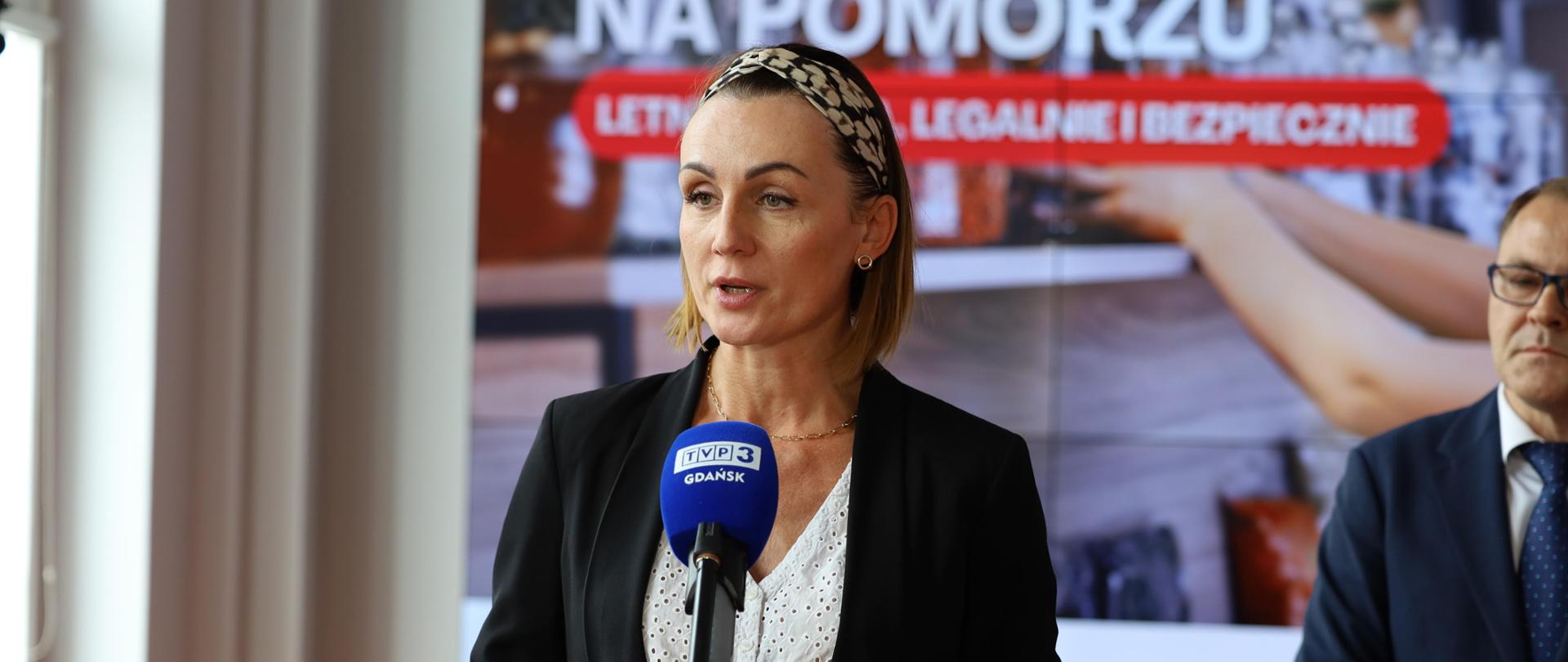 Kobieta - wojewoda pomorska - przemawiająca do mikrofonu podczas konferencji prasowej. Ma na sobie czarną marynarkę, białą bluzkę oraz opaskę na włosy. W tle widoczny jest plakat z napisem "Wakacje na Pomorzu. Letnia praca, legalnie i bezpiecznie". 