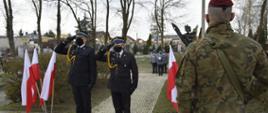 Na pierwszym planie po prawej stronie widzimy stojącego tyłem w pozycji na baczność żołnierza w mundurze moro, berecie z karabinem przewieszonym przez ramie. W dole zdjęcia widać wiązanki kwiatów i stojący na prawo od nich palący się znicz. Przed wiązankami na chodniku wyłożonym kostką stoi dwóch strażaków w mundurach wyjściowych, oddających honor. Obaj mają na twarzach maseczki ochronne. Obok strażaka stojącego z lewej strony w stojaku ustawione są trzy flagi państwowe. Na drugim planie za funkcjonariuszami PSP widać delegację policji. W tle widać pojedyncze wysokie drzewa, krzewy i budynki mieszkalne. 