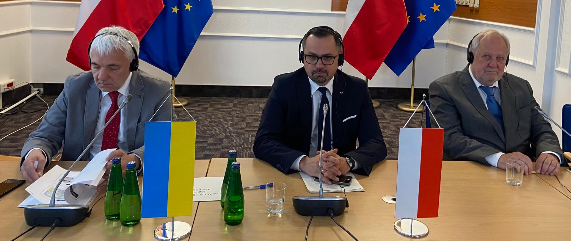 Grupa mężczyzn przy stole, na którym stoją proporczyki Polski i Ukrainy. Za nimi flagi PL i UE.