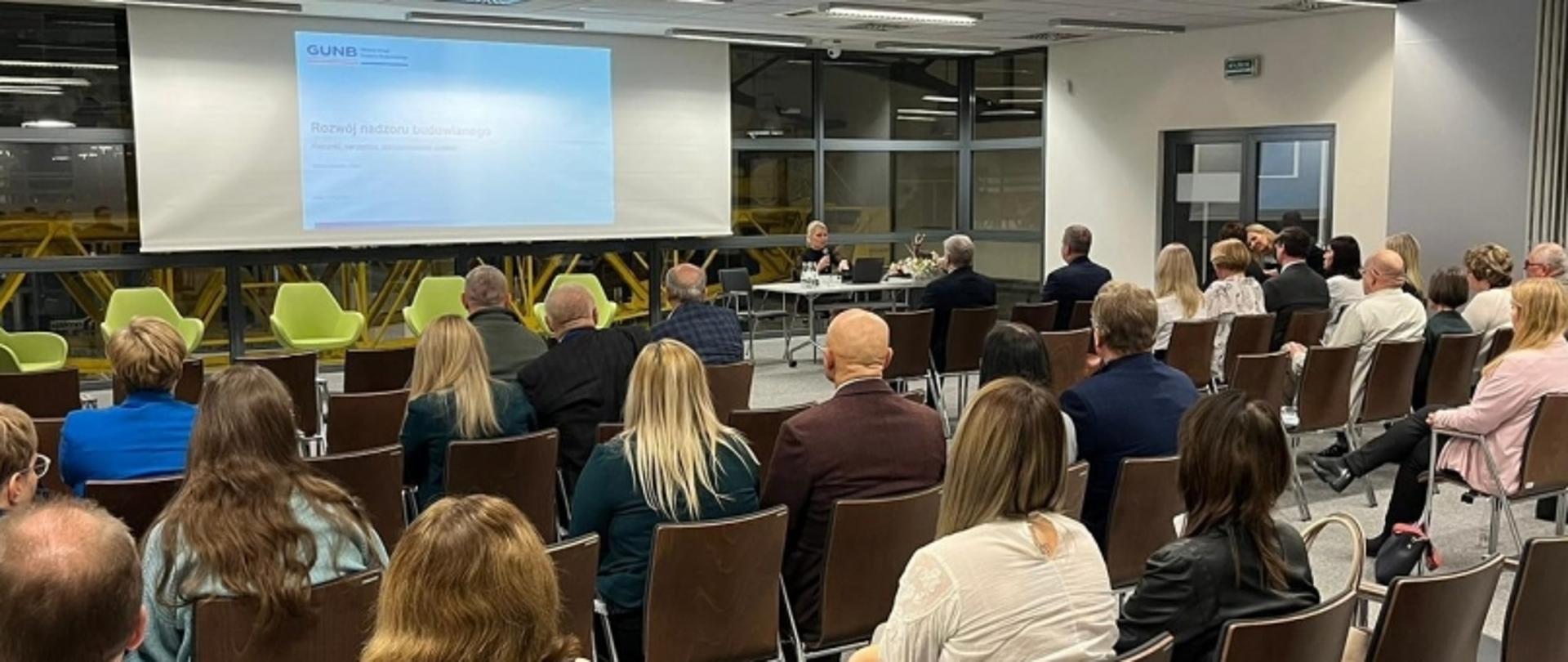Osoby siedzące w sali konferencyjnej, patrzące na ekran z prezentacją „Ruchy kadrowe w budownictwie” i logo GUNB; zielone krzesła i duże okna po lewej stronie.