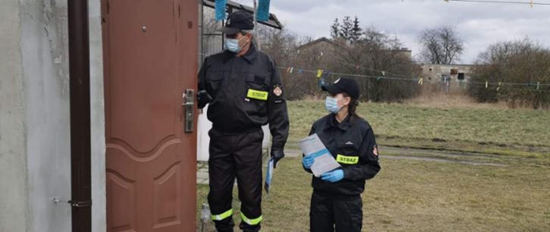 Dwoje druhów OSP w ubraniach koszarowych wręcza ulotki o szczepieniach w mieszkańcowi w otwartych dniach jego domu